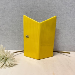 Könnte beinhalten: Eine leuchtend gelbe, rechteckige Vase mit einem strukturierten, vertikal gerippten Design. Die Vase hat eine einzigartige, abgewinkelte Oberseite und ein kleines dekoratives Element auf der Vorderseite. Eine weiße Blume mit langem Stiel liegt neben der Vase.