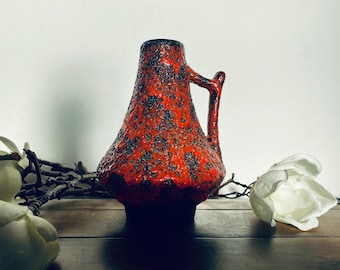 May include: A red and black ceramic vase with a textured surface and a handle. The vase is in a rustic setting with white flowers.