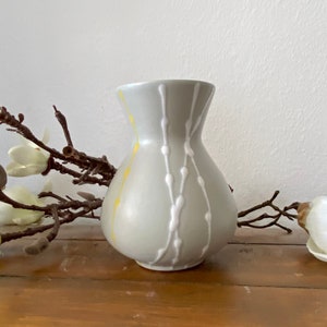 Peut inclure: Un petit vase en céramique gris avec un motif abstrait blanc et jaune. Le vase a un corps arrondi et un col étroit.
