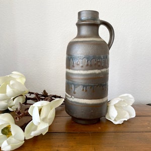 Op de afbeelding: Een bruine en witte keramische vaas met een druppelglazuurontwerp. De vaas heeft een brede opening en een smalle hals. Hij staat op een houten oppervlak met witte bloemen.