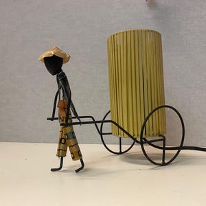 Könnte beinhalten: Eine schwarze Metallfigur eines Menschen, der einen Strohhut trägt und eine Lampe auf einem Wagen transportiert. Der Lampenschirm ist aus geflochtenem Bambus und hat eine gelbe Farbe.