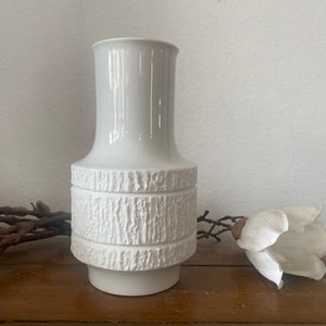 Könnte beinhalten: Weiße Keramikvase mit einem strukturierten, holzähnlichen Design. Die Vase hat eine breite Öffnung und einen schmalen Boden.