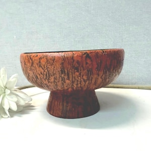 Puede incluir: Un cuenco vintage con pedestal con un exterior texturizado de color marrón rojizo. El cuenco tiene una parte superior ancha y redondeada y una base corta y cilíndrica. Una flor blanca con un tallo largo está a la izquierda del cuenco.