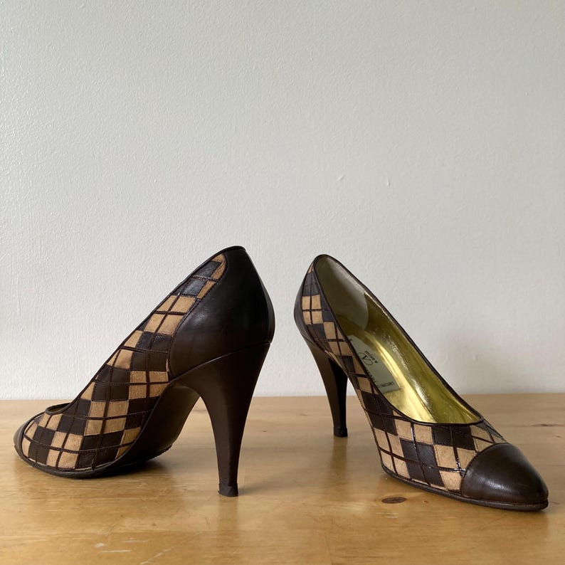 Puede incluir: Un par de zapatos de tac&oacute;n alto marrones y beige. Los zapatos presentan un patr&oacute;n geom&eacute;trico en la parte superior y un tac&oacute;n marr&oacute;n oscuro. La puntera y el tal&oacute;n son de color marr&oacute;n oscuro liso. Los zapatos est&aacute;n sobre una superficie de madera.