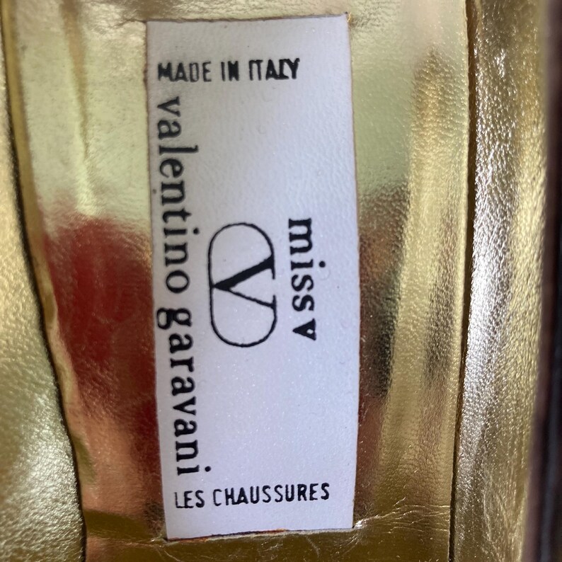 Puede incluir: Primer plano de una etiqueta dentro de un zapato. La etiqueta es blanca con texto negro y un logotipo negro. El texto dice "MADE IN ITALY", "valentino garavani", "miss V" y "LES CHAUSSURES". El interior del zapato es dorado.