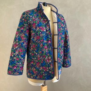 以下が含まれることがあります： 青、ピンク、緑の色合いの鮮やかな花柄のキルティングジャケット。 マンダリンカラー、濃い青色のトリム、ボタン留めが特徴です。 内側の裏地は明るいベージュ色です。 ファッション性の高いアウターウェアです。