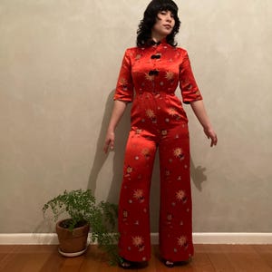 Puede incluir: Un mono de color rojo brillante con cuello mao, mangas tres cuartos y cierres de alamares negros. La prenda está decorada con un patrón repetitivo de motivos florales dorados y blancos y tiene pantalones anchos de corte recto.