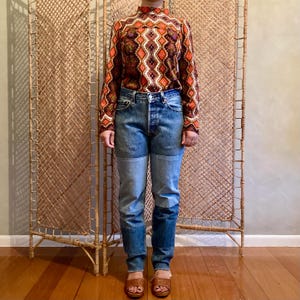 May include: A person wearing a long-sleeved, high-neck patterned top in shades of brown, orange, and white, paired with blue denim jeans featuring patchwork details. The outfit is completed with brown sandals. The background includes a woven screen.