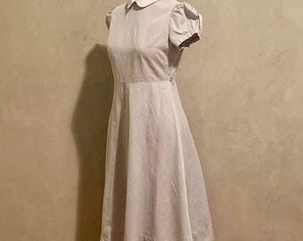 Może przedstawiać: Sukienka w stylu vintage prezentowana na manekinie. Sukienka posiada biały kołnierzyk, krótkie bufiaste rękawy i dopasowaną talię, która przechodzi w rozkloszowaną spódnicę. Tkanina ma subtelny wzór w paski w jasnym kolorze.