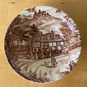 Puede incluir: Un plato redondo de cerámica marrón y blanco con una ilustración detallada de una escena de pueblo. El diseño incluye edificios, un carruaje tirado por caballos y figuras, con un castillo en una colina en el fondo. El plato tiene un acabado brillante.