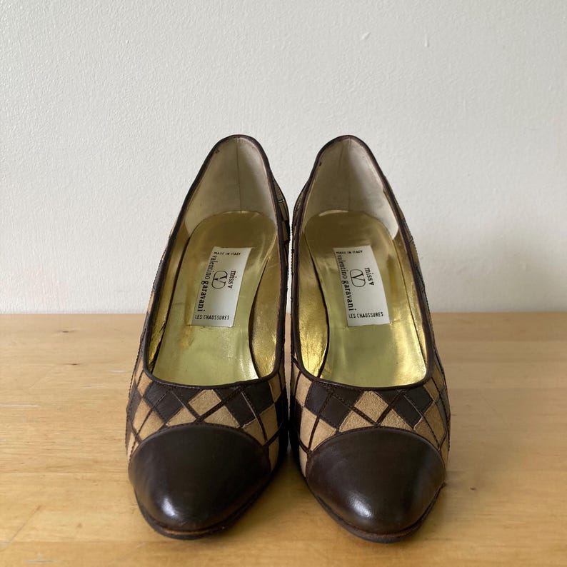 Puede incluir: Un par de zapatos de tac&oacute;n alto marrones y beige con un patr&oacute;n de diamantes en los laterales. La puntera y el tal&oacute;n son de color marr&oacute;n liso. El interior es dorado y la etiqueta dice "Valentino Garavani".
