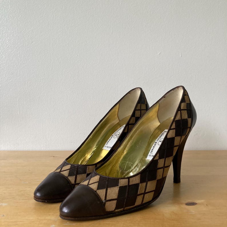 Puede incluir: Un par de zapatos de tac&oacute;n alto marrones y beige con un patr&oacute;n de diamantes. Los zapatos tienen una punta puntiaguda y un tac&oacute;n de aguja. El interior es dorado. Los zapatos est&aacute;n sobre una superficie de madera.