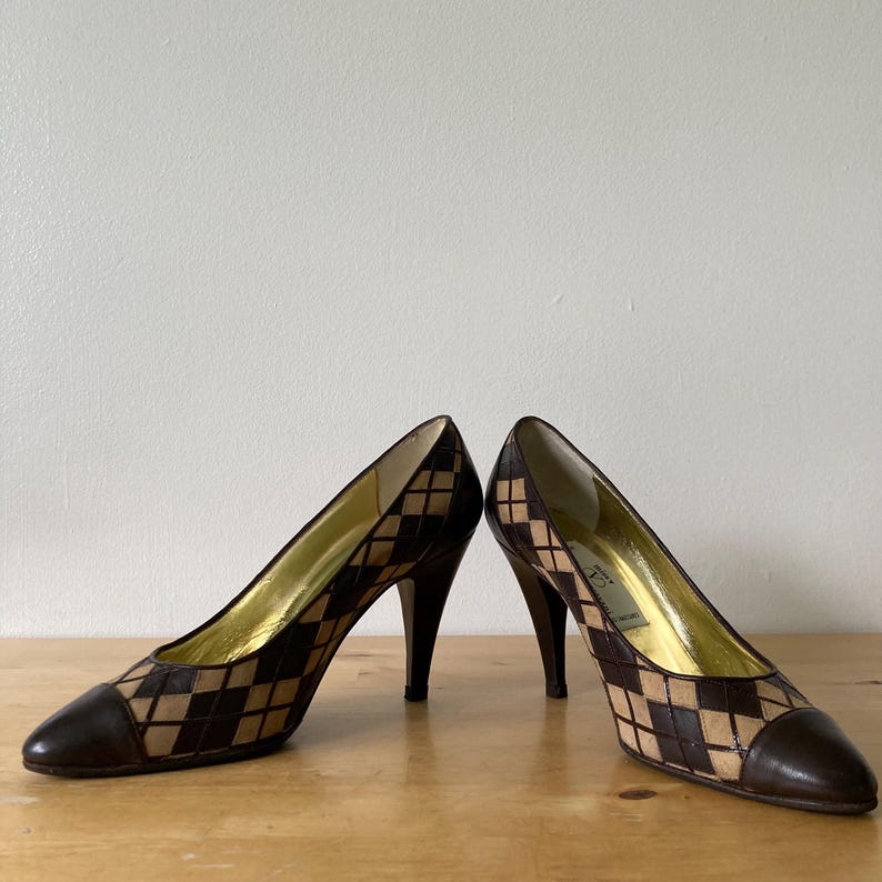 Puede incluir: Un par de zapatos de tac&oacute;n alto con un patr&oacute;n de diamantes marr&oacute;n y beige. Los zapatos tienen una punta puntiaguda y un tac&oacute;n de aguja. El interior del zapato es dorado.