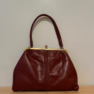May include: A dark red leather handbag featuring a structured gold-tone metal frame, a single arched handle, and a central vertical seam detail. The bag has a kiss-lock closure at the top center and a soft, slightly creased texture throughout.