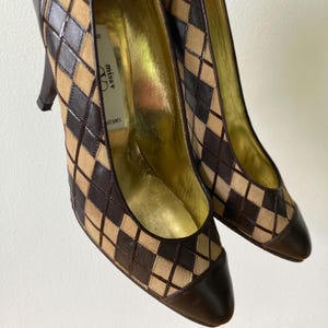 Puede incluir: Un par de zapatos de tac&oacute;n alto con un patr&oacute;n de diamantes marr&oacute;n y beige. Los zapatos tienen una punta puntiaguda y un tac&oacute;n marr&oacute;n oscuro. El interior es dorado y la marca "miss v" es visible en la plantilla.