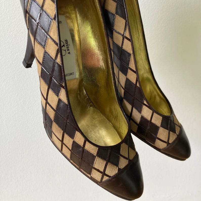 Puede incluir: Un par de zapatos de tac&oacute;n alto con un patr&oacute;n de diamantes marr&oacute;n y beige. Los zapatos tienen una punta puntiaguda y un tac&oacute;n marr&oacute;n oscuro. El interior es dorado y la marca "miss v" es visible en la plantilla.