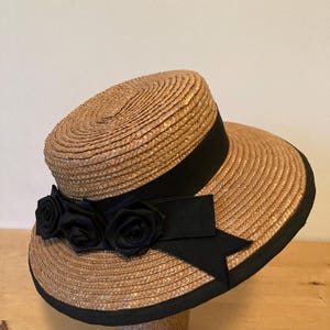 May include: A straw hat with a wide brim and a black grosgrain ribbon. The hat features three black fabric roses and a black trim along the edge. The hat is designed for sun protection and fashion.