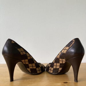 Puede incluir: Un par de zapatos de tac&oacute;n marr&oacute;n. Los tacones y la parte trasera de los zapatos son de color marr&oacute;n oscuro liso. La parte delantera de los zapatos tiene un estampado de cuadros beige y marr&oacute;n. Los zapatos est&aacute;n sobre una superficie de madera.