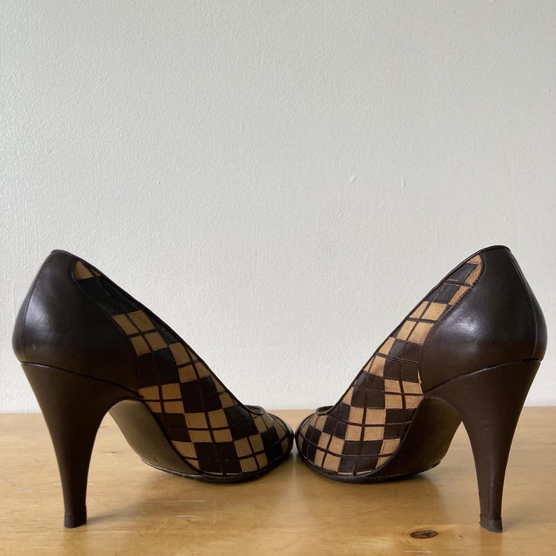 Puede incluir: Un par de zapatos de tac&oacute;n marr&oacute;n. Los tacones y la parte trasera de los zapatos son de color marr&oacute;n oscuro liso. La parte delantera de los zapatos tiene un estampado de cuadros beige y marr&oacute;n. Los zapatos est&aacute;n sobre una superficie de madera.