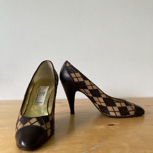 Puede incluir: Un par de zapatos de tac&oacute;n alto marrones y beige con un patr&oacute;n de diamantes. Los zapatos tienen una punta afilada y un tac&oacute;n de aguja. El interior del zapato es dorado. Los zapatos est&aacute;n sobre una superficie de madera.