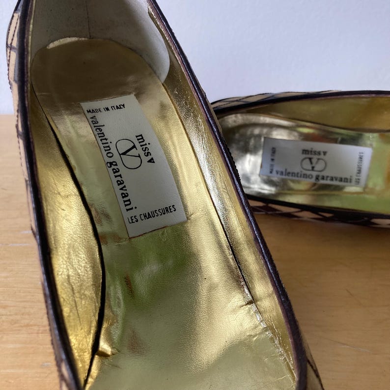 Puede incluir: Primer plano de un par de tacones Valentino Garavani. El interior es dorado con una etiqueta que dice "miss V" y "valentino garavani LES CHAUSSURES". El exterior tiene un estampado con formas marrones y beige.
