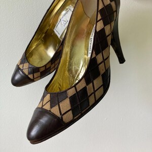 Puede incluir: Un par de tacones Valentino Garavani. Los zapatos presentan un patr&oacute;n de diamantes marr&oacute;n y beige en la parte superior, con una puntera y un tal&oacute;n de cuero marr&oacute;n oscuro. El interior es dorado y los tacones miden unos 7.6 cm de alto.