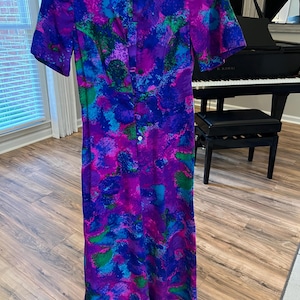 Puede incluir: Vestido vintage largo hasta el suelo con un vibrante estampado abstracto en tonos morado, azul y verde. El vestido presenta mangas cortas, una botonadura frontal y una pequeña etiqueta en el escote. El vestido está hecho de tela ligera.