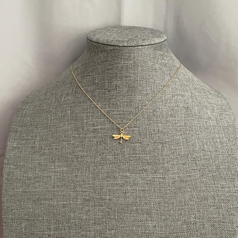 May include: A gold chain necklace with a delicate dragonfly pendant. The necklace is displayed on a gray fabric mannequin.