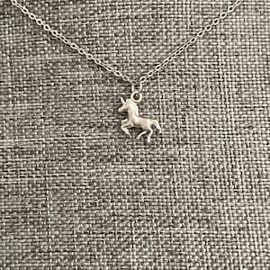 Könnte beinhalten: Eine silberne Halskette mit einem Einhorn-Anhänger. Die Halskette liegt auf einem grauen, strukturierten Hintergrund.