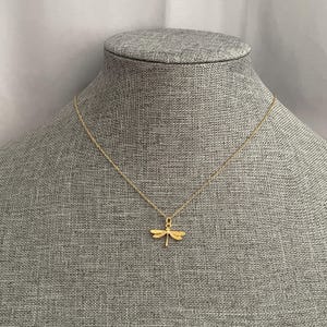May include: A gold chain necklace with a delicate dragonfly pendant. The necklace is displayed on a gray fabric mannequin.