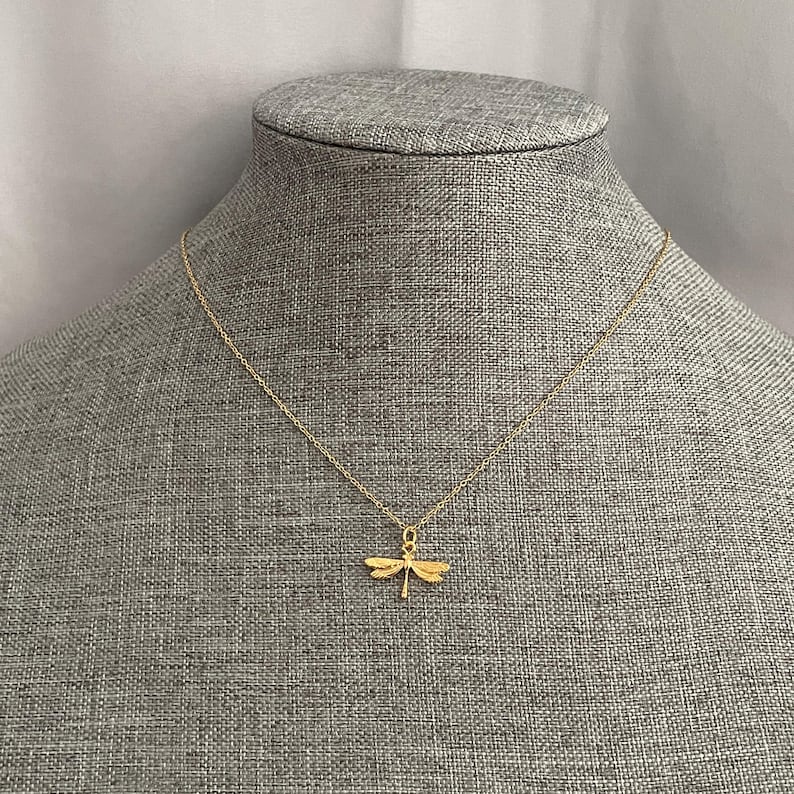 May include: A gold chain necklace with a delicate dragonfly pendant. The necklace is displayed on a gray fabric mannequin.