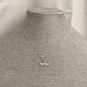 May include: A silver necklace with a dragonfly pendant. The necklace is on a gray fabric background.