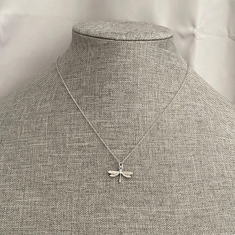 May include: A silver necklace with a dragonfly pendant. The necklace is on a gray fabric background.