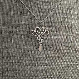 Puede incluir: Collar plateado con un colgante calado ornamentado. El colgante presenta un diseño simétrico con patrones arremolinados y un pequeño dije de hoja. La cadena es delicada y el fondo es gris. Una pieza de joyería.
