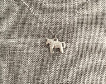 May include: A silver-toned necklace featuring a pendant in the shape of a Dala horse. The horse pendant has detailed etched designs on its body and is suspended from a delicate chain. The necklace is displayed against a textured gray background.