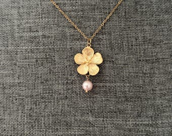 May include: A gold-toned necklace featuring a delicate chain and a five-petaled flower pendant with a textured surface. A single, small, round, pale pink bead is suspended directly beneath the center of the gold flower charm.