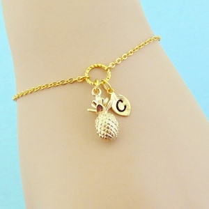 May include: A gold chain bracelet with a pineapple charm and a letter 'C' charm. The bracelet is on a light blue background.