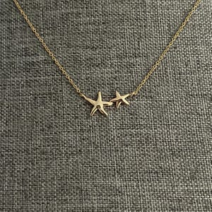 Peut inclure: Un collier doré composé de deux pendentifs en forme d'étoile de mer de tailles différentes. La chaîne fine est fixée aux extrémités de la plus grande étoile, tandis que la plus petite est placée juste à côté, formant un ensemble harmonieux.