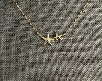 May include: A gold-toned necklace featuring two connected starfish charms of different sizes. The delicate chain is attached to the sides of the larger starfish, while the smaller starfish is positioned directly next to it, creating a cohesive design.