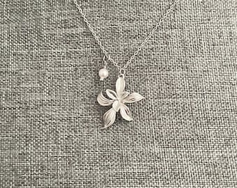 May include: A silver necklace with a delicate flower pendant and a single pearl charm. The necklace is on a gray textured background.
