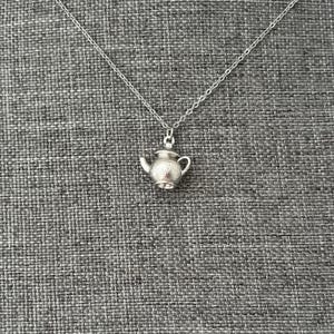 May include: A silver necklace featuring a miniature teapot pendant. The teapot charm is decorated with floral patterns and has a handle. The chain is delicate, set against a grey textured backdrop.