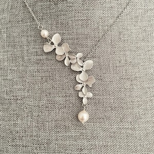 Peut inclure: Un collier argenté avec un pendentif floral. Le pendentif présente cinq grappes de fleurs et deux perles. Le collier a une chaîne délicate et une perle en bas. Les fleurs ont une finition texturée.