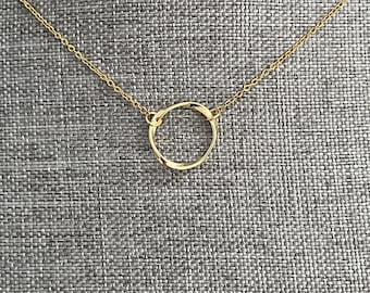 May include: A delicate gold-colored necklace featuring a small, open circle pendant. The pendant has a textured surface and is suspended from a fine chain. The necklace is displayed against a gray textured background.