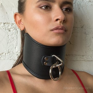 A woman with her brown hair in a ponytail is shown from the shoulders up standing in front of a white brick wall. She is wearing the Tall Curved Posture Collar With A Locking Buckle. The collar covers her entire neck, from her clavicle to her chin.