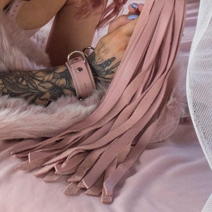 A close-up of a  tattooed woman's forearms, lying on a pink bed, is shown. She holds the Stupid Cute Baby Pink Leather Flogger. The handle is obscured by the many light pink falls which drape onto the bed.