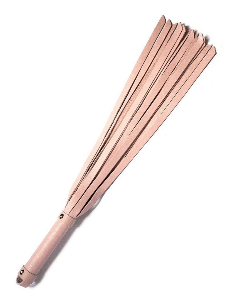 The Stupid Cute Baby Pink Leather Flogger is shown against a blank background. The handle is smooth, cylindrical, and the same color leather as the falls. There is a small loop at the base of the handle.