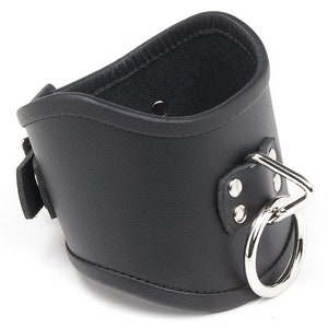 The Tall Curved Posture Collar With A Locking Buckle is shown against a blank background. It is black leather with silver hardware and has a D-ring in the front with an O-ring hanging off of it. The collar has a slight dip at the top in the front.