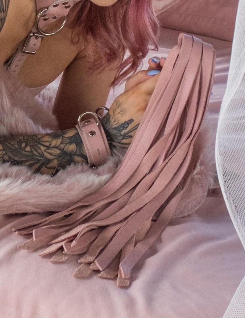 A close-up of a  tattooed woman's forearms, lying on a pink bed, is shown. She holds the Stupid Cute Baby Pink Leather Flogger. The handle is obscured by the many light pink falls which drape onto the bed.