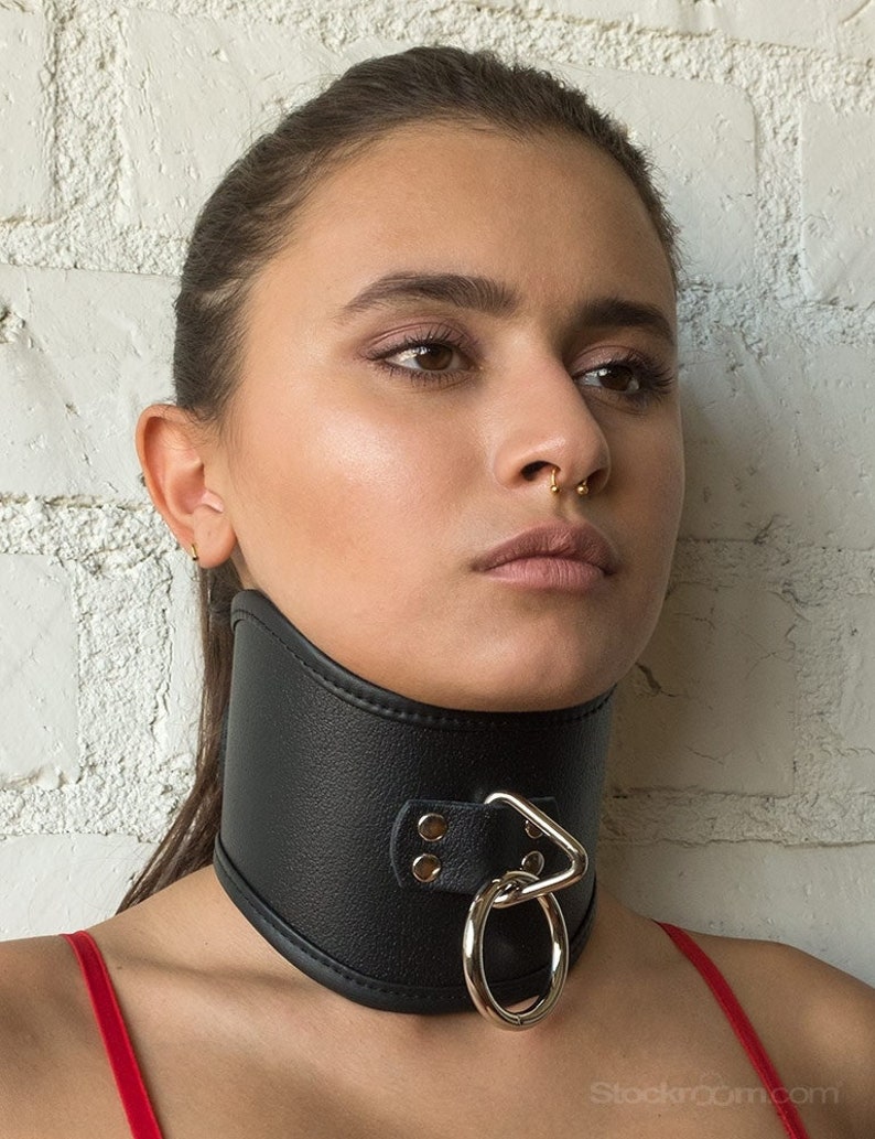 A woman with her brown hair in a ponytail is shown from the shoulders up standing in front of a white brick wall. She is wearing the Tall Curved Posture Collar With A Locking Buckle. The collar covers her entire neck, from her clavicle to her chin.