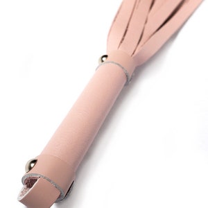 A close-up of the handle of the Stupid Cute Baby Pink Leather Flogger is shown against a blank background, displaying the small leather loop at the base of the handle and the metal hardware.